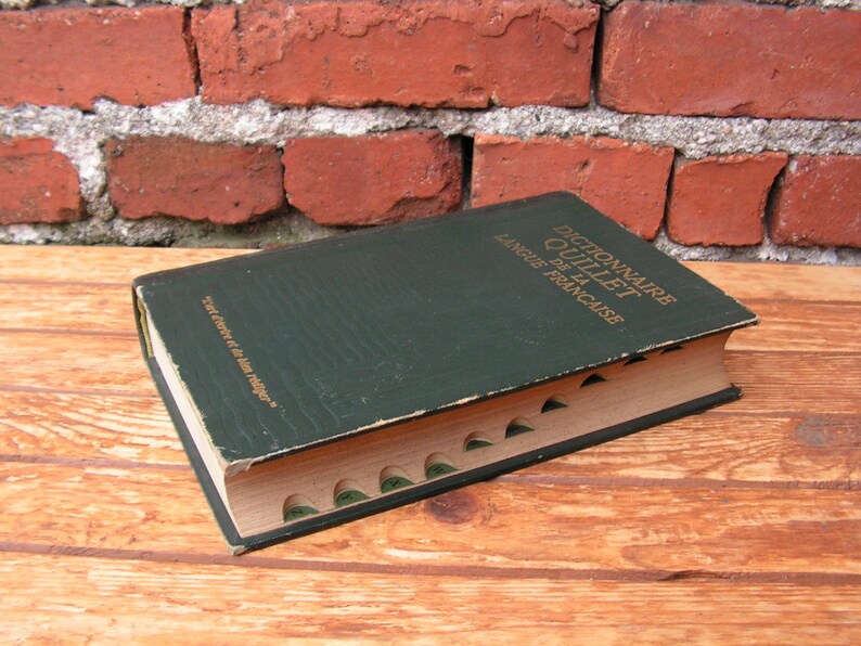 May include: A vintage green hardcover book titled "Dictionnaire Quillet de la Langue Fran&ccedil;aise" with gold lettering. The book has a worn appearance and is open to show the interior pages. The book is on a wooden surface.