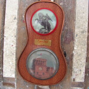 May include: A wooden plaque with two round glass inserts. The top insert shows a black and white image of a rider on horseback. The bottom insert shows a black and white image of a building. The plaque has a gold label with the text "Erebuni 1970".