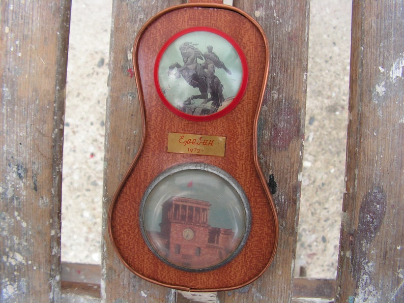 May include: A wooden plaque with two round glass inserts. The top insert shows a black and white image of a rider on horseback. The bottom insert shows a black and white image of a building. The plaque has a gold label with the text "Erebuni 1970".