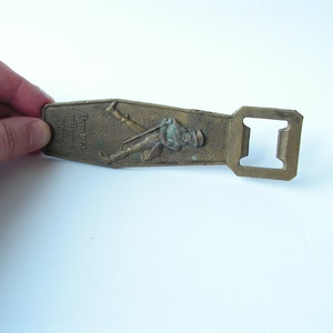 May include: Vintage brass bottle opener with a rectangular shape and a built-in bottle cap remover. The opener features an embossed image of a man in a hat and the text "Born 1850 still going strong".