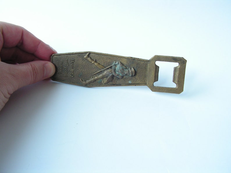 May include: Vintage brass bottle opener with a rectangular shape and a built-in bottle cap remover. The opener features an embossed image of a man in a hat and the text "Born 1850 still going strong".