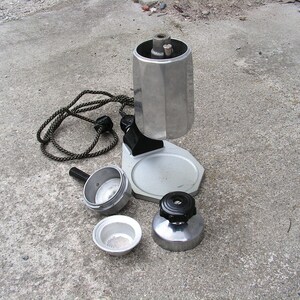 May include: A silver and black electric stovetop espresso maker with a removable filter basket and a cord. The maker is disassembled and laid out on a gray concrete surface.