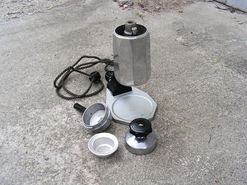 May include: A silver and black electric stovetop espresso maker with a removable filter basket and a cord. The maker is disassembled and laid out on a gray concrete surface.