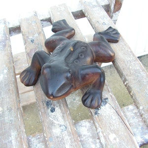 May include: A dark brown ceramic frog figurine with a textured surface. The frog is sitting on a wooden surface with white paint splatters.