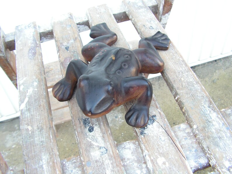May include: A dark brown ceramic frog figurine with a textured surface. The frog is sitting on a wooden surface with white paint splatters.