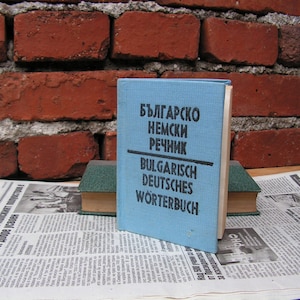 Op de afbeelding: Een blauw hardback boek met de titel "Bulgarsko Nemski Rechnik" en "Bulgarisch Deutsches Wörterbuch" gedrukt op de omslag. Het boek ligt op een stapel andere boeken.