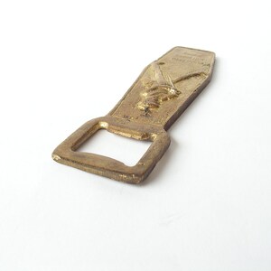 May include: A gold-coloured bottle opener with a raised design of two crossed swords. The opener has a rectangular shape with a rounded top and a small opening for prying off bottle caps.