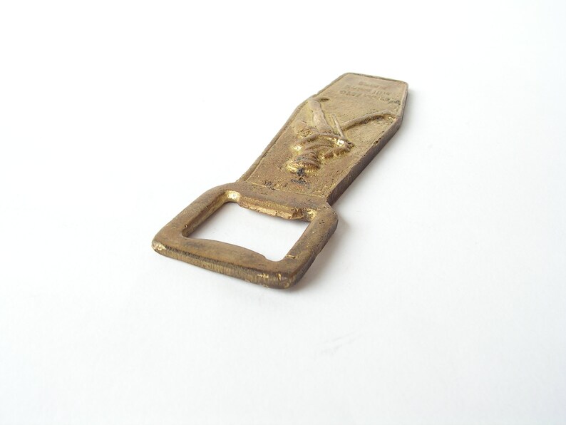 May include: A gold-coloured bottle opener with a raised design of two crossed swords. The opener has a rectangular shape with a rounded top and a small opening for prying off bottle caps.