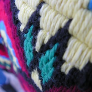 May include: Close-up of a colorful, textured textile with a geometric pattern. The design features yellow, black, teal, and magenta yarn, creating a vibrant and detailed surface. The knitted or crocheted fabric has a handmade appearance.
