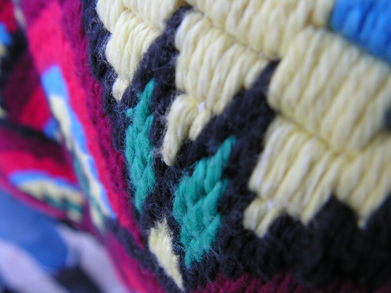 May include: Close-up of a colorful, textured textile with a geometric pattern. The design features yellow, black, teal, and magenta yarn, creating a vibrant and detailed surface. The knitted or crocheted fabric has a handmade appearance.