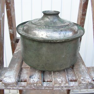 May include: A vintage, silver-colored metal pot with a lid, sitting on a wooden step ladder. The pot has a green patina and is slightly tarnished.