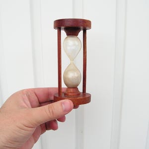 Könnte beinhalten: Eine hölzerne Sanduhr mit dunkelbraunem Finish. Die Sanduhr hat eine klare Glaskugel, die mit hellfarbenem Sand gefüllt ist. Die Sanduhr ist etwa 10 cm hoch.