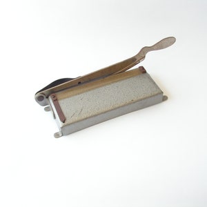 May include: A vintage metal paper cutter with a gray base and a silver handle. The cutter has a small metal blade and a brown plastic guide.