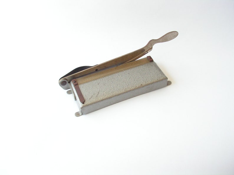 May include: A vintage metal paper cutter with a gray base and a silver handle. The cutter has a small metal blade and a brown plastic guide.
