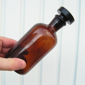 May include: A brown glass bottle with a black plastic stopper. The bottle is empty and has a smooth, shiny surface.