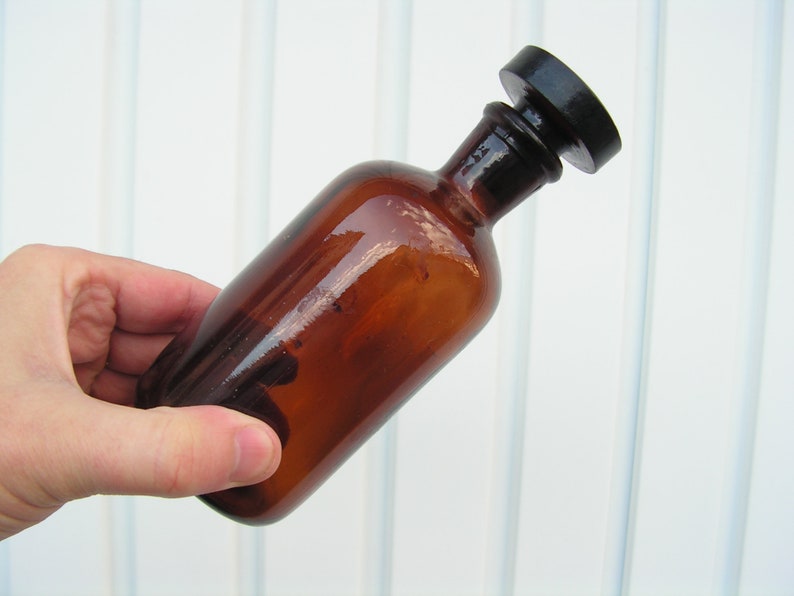 May include: A brown glass bottle with a black plastic stopper. The bottle is empty and has a smooth, shiny surface.