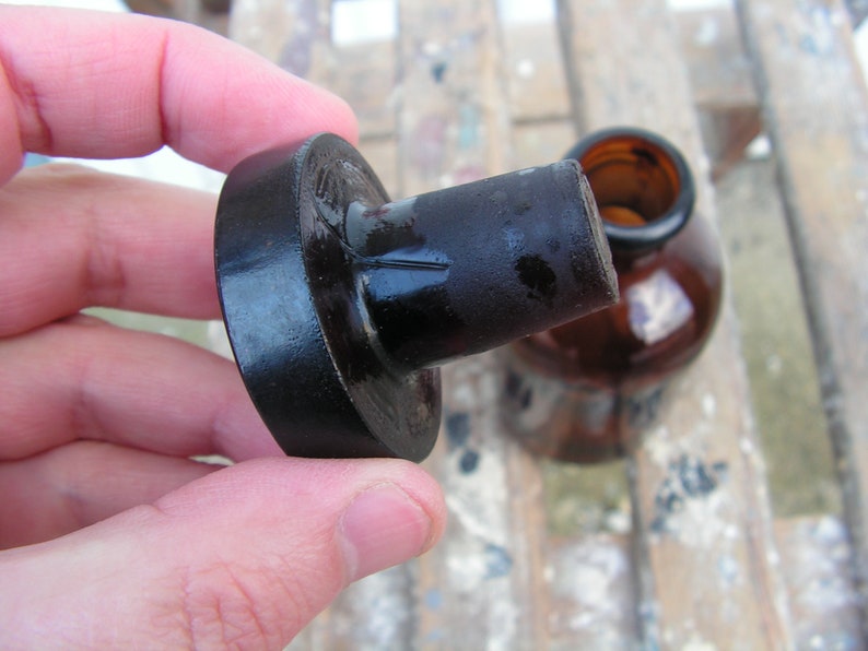 May include: A black glass bottle stopper with a round, flat top. The stopper is made of dark glass and has a smooth, shiny finish.