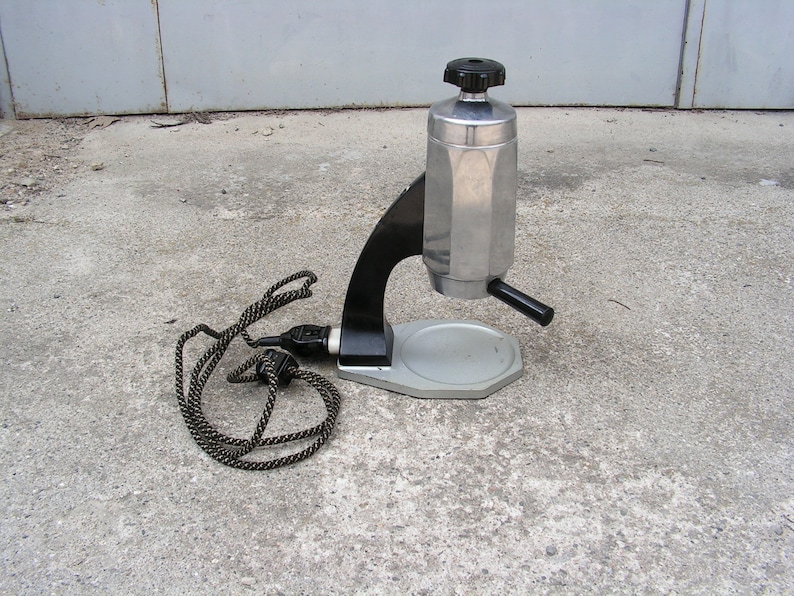 May include: A silver and black electric coffee maker with a cord and plug. The coffee maker has a round base and a tall, cylindrical pot with a black handle.