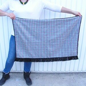 May include: A rectangular plaid blanket with a black fringe along the bottom edge. The plaid pattern features blue, red, and white lines on a navy background. The blanket is held up by a person.