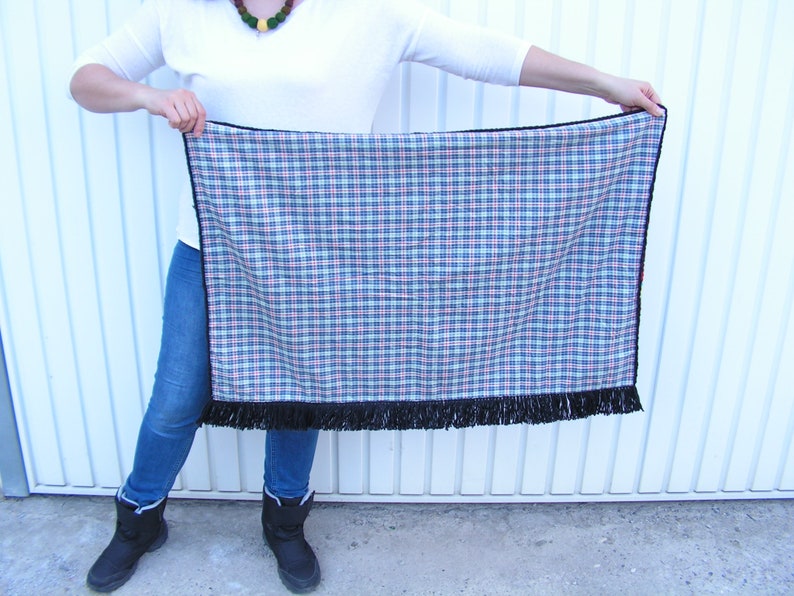 May include: A rectangular plaid blanket with a black fringe along the bottom edge. The plaid pattern features blue, red, and white lines on a navy background. The blanket is held up by a person.