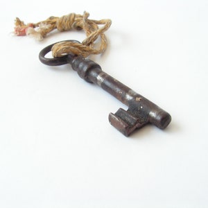 May include: An antique, rusty metal key with a circular handle and a rectangular bit. A twisted, beige rope is attached to the key's handle. The key is lying on a white surface.