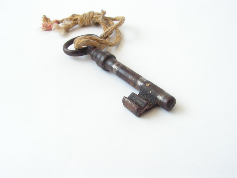 May include: An antique, rusty metal key with a circular handle and a rectangular bit. A twisted, beige rope is attached to the key's handle. The key is lying on a white surface.