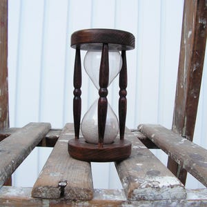 May include: A wooden hourglass with a clear glass bulb and white sand. The hourglass is sitting on a wooden shelf.