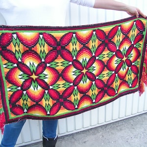 May include: A rectangular, woven textile with a vibrant floral pattern in red, orange, yellow, and green. The design features repeating flower-like shapes with a black outline. The edges are trimmed with a green border and red fringe.