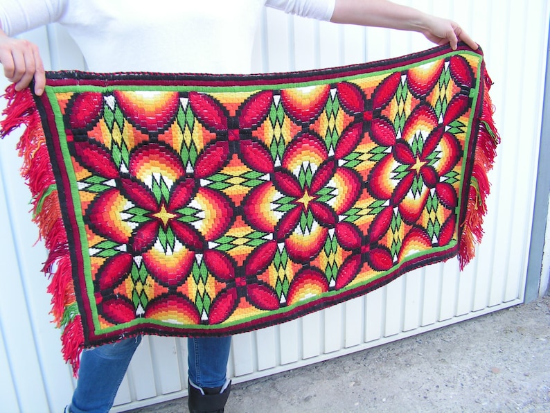 May include: A rectangular, woven textile with a vibrant floral pattern in red, orange, yellow, and green. The design features repeating flower-like shapes with a black outline. The edges are trimmed with a green border and red fringe.