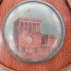 May include: A round, glass-covered image of a building with a clock tower. The building is painted in shades of brown and red, with a white flag on top. The image is set in a circular, metal frame.