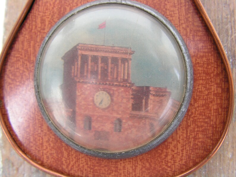 May include: A round, glass-covered image of a building with a clock tower. The building is painted in shades of brown and red, with a white flag on top. The image is set in a circular, metal frame.