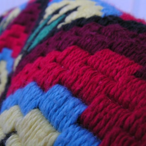May include: Close-up of a colorful, textured textile with a geometric pattern. The design features vibrant red, blue, yellow, and green yarn, creating a visually appealing and intricate design. The fabric appears to be hand-stitched or crocheted, showcasing a handcrafted aesthetic.
