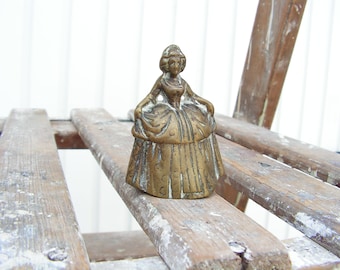 Op de afbeelding: Een messing bel in de vorm van een vrouw in een lange jurk. De bel staat op een houten oppervlak.