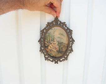 May include: An oval-shaped picture frame with an ornate, gold-toned metal design. The frame holds a vintage print of a young girl in a garden setting.