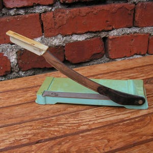 May include: A vintage paper cutter with a white handle and a green base. The cutter has a black blade and is sitting on a wooden surface.