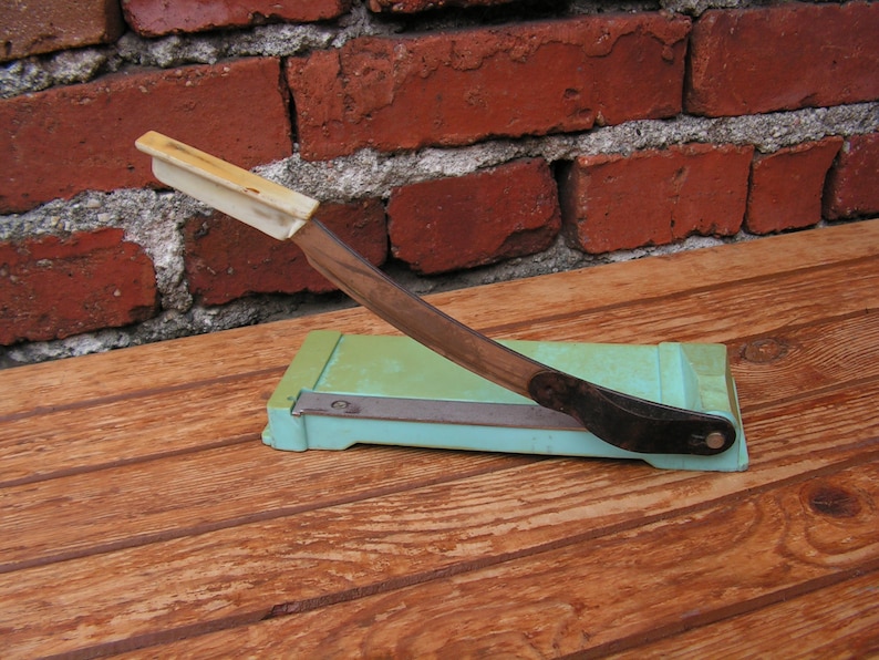 May include: A vintage paper cutter with a white handle and a green base. The cutter has a black blade and is sitting on a wooden surface.