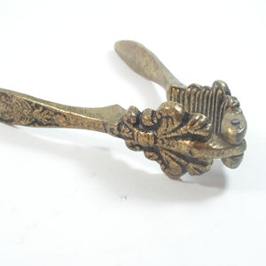 Puede incluir: Un cascanueces de metal dorado con diseños florales ornamentados en las asas. El cascanueces tiene un área estriada y texturizada para romper nueces.