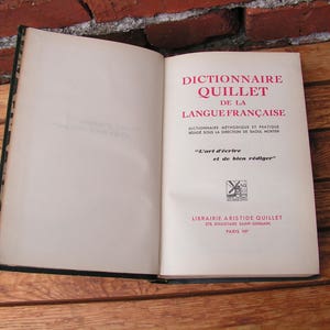May include: An open book with a black cover and aged pages. The title page reads "Dictionnaire Quillet de la Langue Fran&ccedil;aise" in red and black text. The book is on a wooden surface.