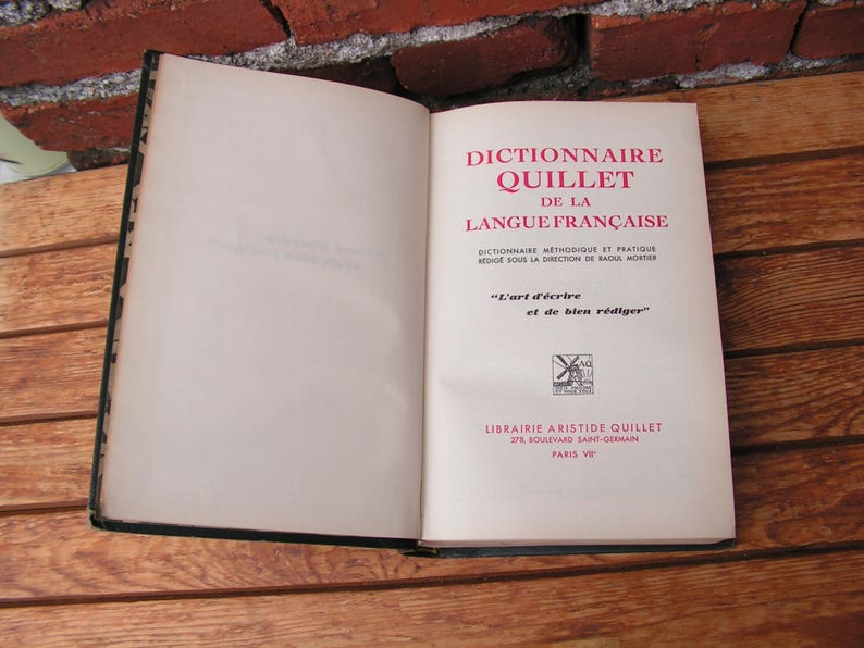 May include: An open book with a black cover and aged pages. The title page reads "Dictionnaire Quillet de la Langue Fran&ccedil;aise" in red and black text. The book is on a wooden surface.