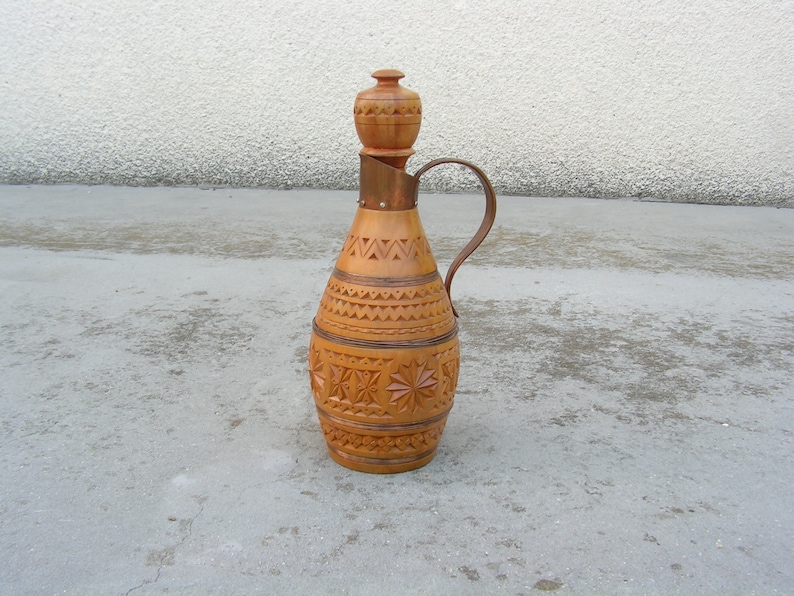 K&ouml;nnte beinhalten: Eine Holzflasche mit einem geschnitzten Design und einem kupfernen Oberteil und Griff. Die Flasche ist braun und hat einen traditionellen Volksstil.
