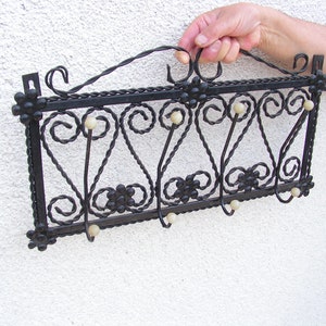 Puede incluir: Perchero de pared de metal negro con decoración de volutas y pomos de plástico blanco. El perchero tiene cinco ganchos.