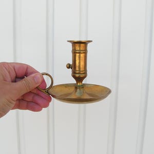 Könnte beinhalten: Ein Vintage-Kerzenhalter aus Messing mit rundem Sockel und Griff. Der Kerzenhalter hat ein klassisches Design mit einer Tasse zum Halten einer Kerze. Das Messing hat eine warme, gealterte Patina. Der Halter ist ca. 13 cm hoch.