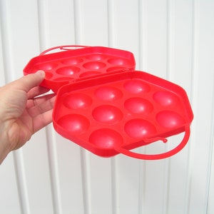 May include: A red plastic egg carrier with a handle. The carrier has 12 individual compartments for holding eggs.