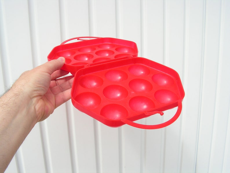 May include: A red plastic egg carrier with a handle. The carrier has 12 individual compartments for holding eggs.