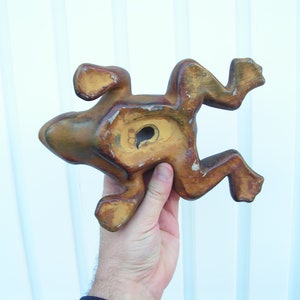 May include: A wooden frog figurine with a brown and yellow paint finish. The frog is in a jumping pose with its legs outstretched. The frog has a hole in its back, suggesting it may be a wall hanging.