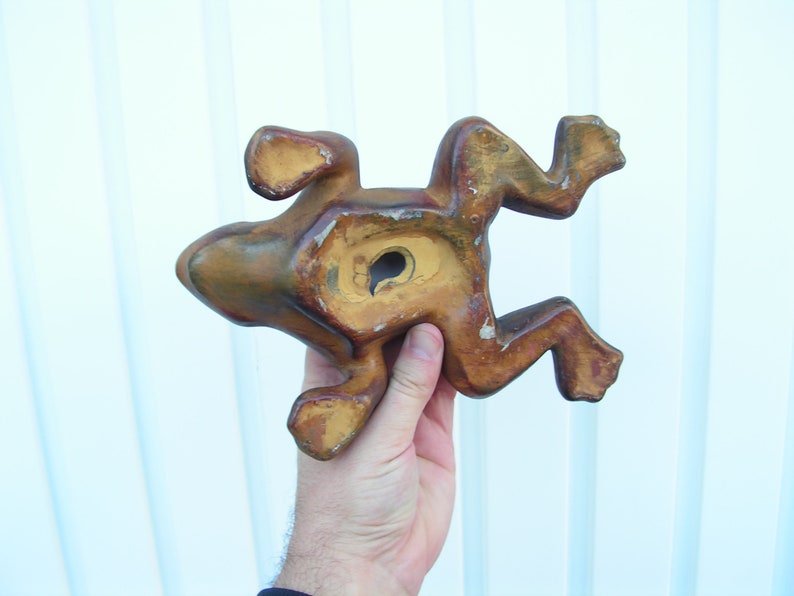 May include: A wooden frog figurine with a brown and yellow paint finish. The frog is in a jumping pose with its legs outstretched. The frog has a hole in its back, suggesting it may be a wall hanging.