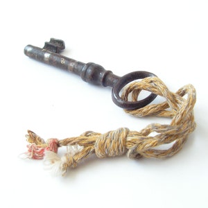 May include: An antique metal key with a circular handle is tied with a beige and tan rope. The key is dark brown with a worn appearance. The rope has a knotted design with frayed ends, and a touch of red thread.