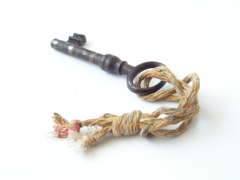 May include: An antique metal key with a circular handle is tied with a beige and tan rope. The key is dark brown with a worn appearance. The rope has a knotted design with frayed ends, and a touch of red thread.