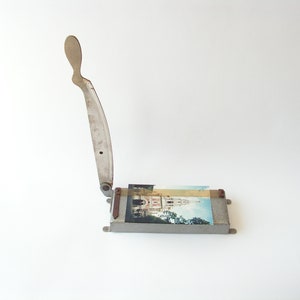May include: A vintage metal paper cutter with a silver blade and a handle. The cutter is sitting on a white surface and has a photo of a building in the background.