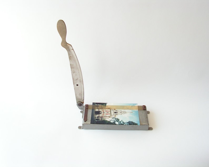 May include: A vintage metal paper cutter with a silver blade and a handle. The cutter is sitting on a white surface and has a photo of a building in the background.
